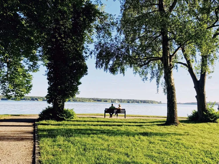 Zwei Menschen auf einer Parkbank unter großen Bäumen am Seeufer, auf dem Wasser ein Segelboot