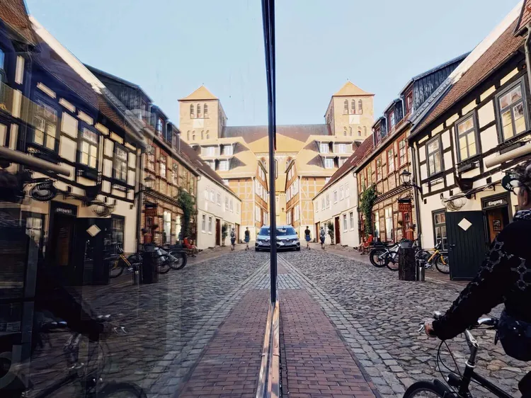 Street-Fotografie: Altstadtgasse mit Fachwerkhäusern und zwei Backsteintürmen, symmetrisch in einer Schaufensterscheibe gespiegelt, rechts ein Radfahrer