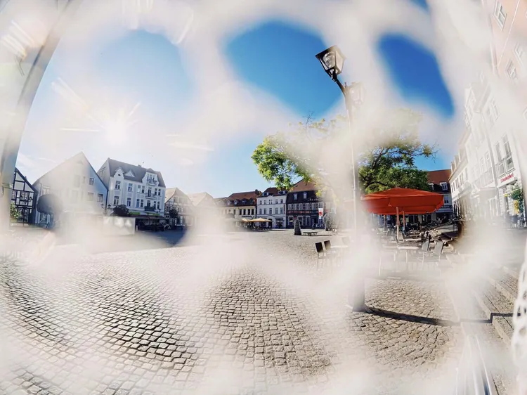 Sonniger Marktplatz mit Kopfsteinpflaster, Fachwerkhäusern und rotem Sonnenschirm, fotografiert durch die Maschen eines Netzes