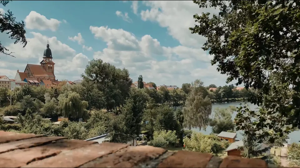 Blick über Gärten und Bäume auf die Altstadt von Waren (Müritz) mit Kirchturm, Seeufer und Sommerwolken – Vorschaubild für das TimeLapse
